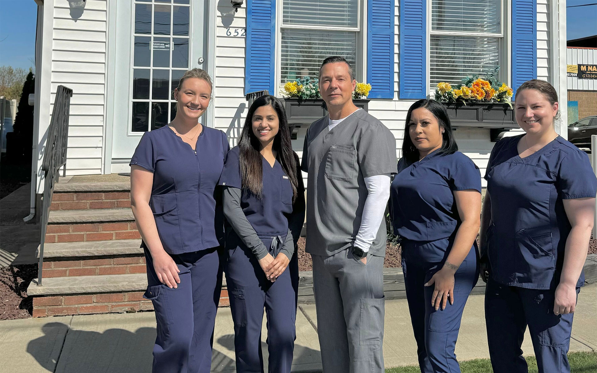 A group of five people, likely nurses due to their uniforms, posing for a photo outside a building with a blue and white striped awning.