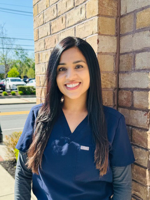 A woman wearing scrubs stands outside a brick building with a smile on her face.