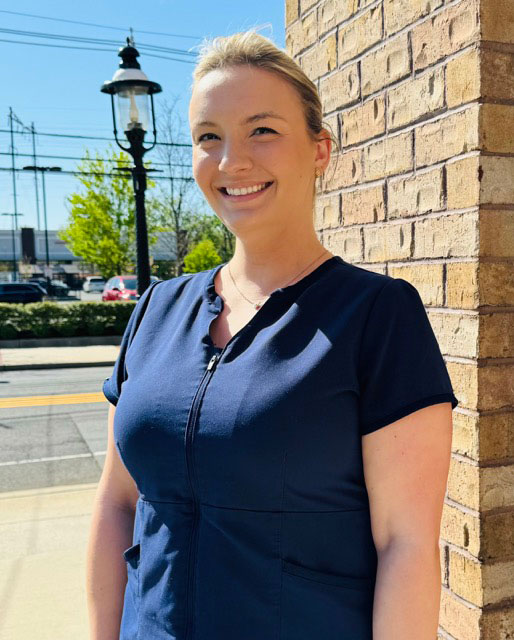 The image shows a woman standing outdoors in front of a brick building, smiling at the camera.