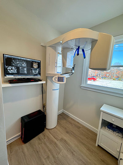 The image shows an interior space with medical equipment, including a large dental chair and a digital X-ray machine, set against a backdrop of white walls and a window allowing natural light into the room.