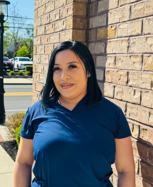 The image shows a person standing outdoors, wearing a dark blue shirt and black pants, with short hair, smiling at the camera.