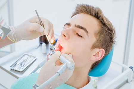 A young man undergoing dental treatment, with a dentist using a drill on his teeth.
