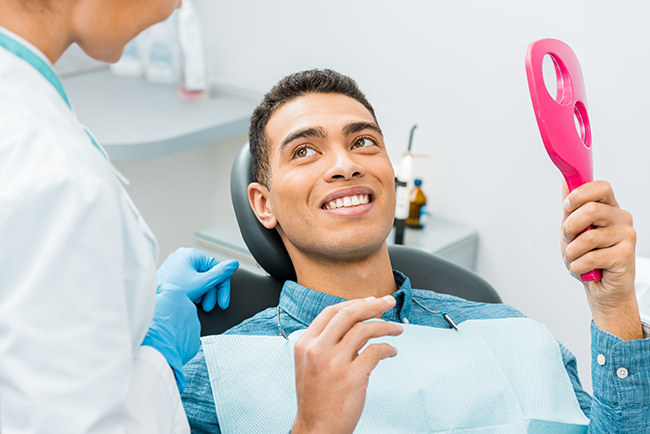 The image shows a person sitting in a dental chair with a smile, holding a pink object that resembles a toothbrush, while a dental professional attends to them.