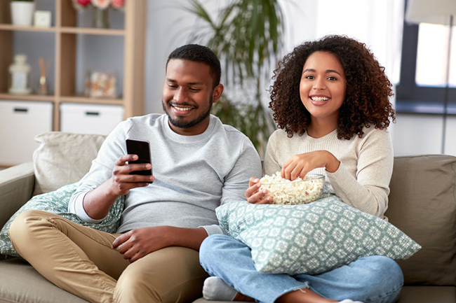 A man and woman sitting on a couch, smiling at each other while holding snacks, with a cell phone between them displaying content.